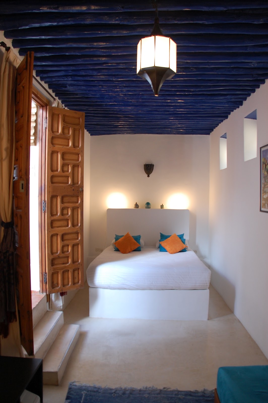 Chambre Zerka : plafond de poutres peintes en bleu, porte en cèdre sculpté ouverte et lit aux coussins orange et turquoise