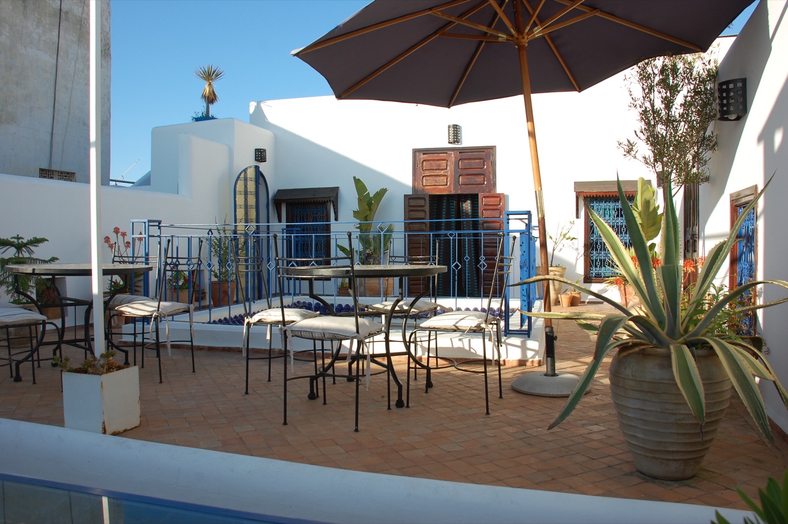 La terrasse du riad : tables en fer forgé, parasol, plantes et murs blanchis à la chaux sous le ciel bleu